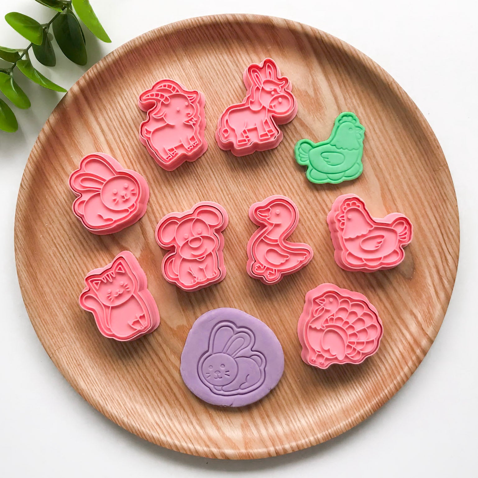 A set of pink animal-shaped cutters on a wooden surface, with a variety of animal figures such as rabbits, chickens, and ducks. There is also a wooden cutter piece with a design of a rabbit.
