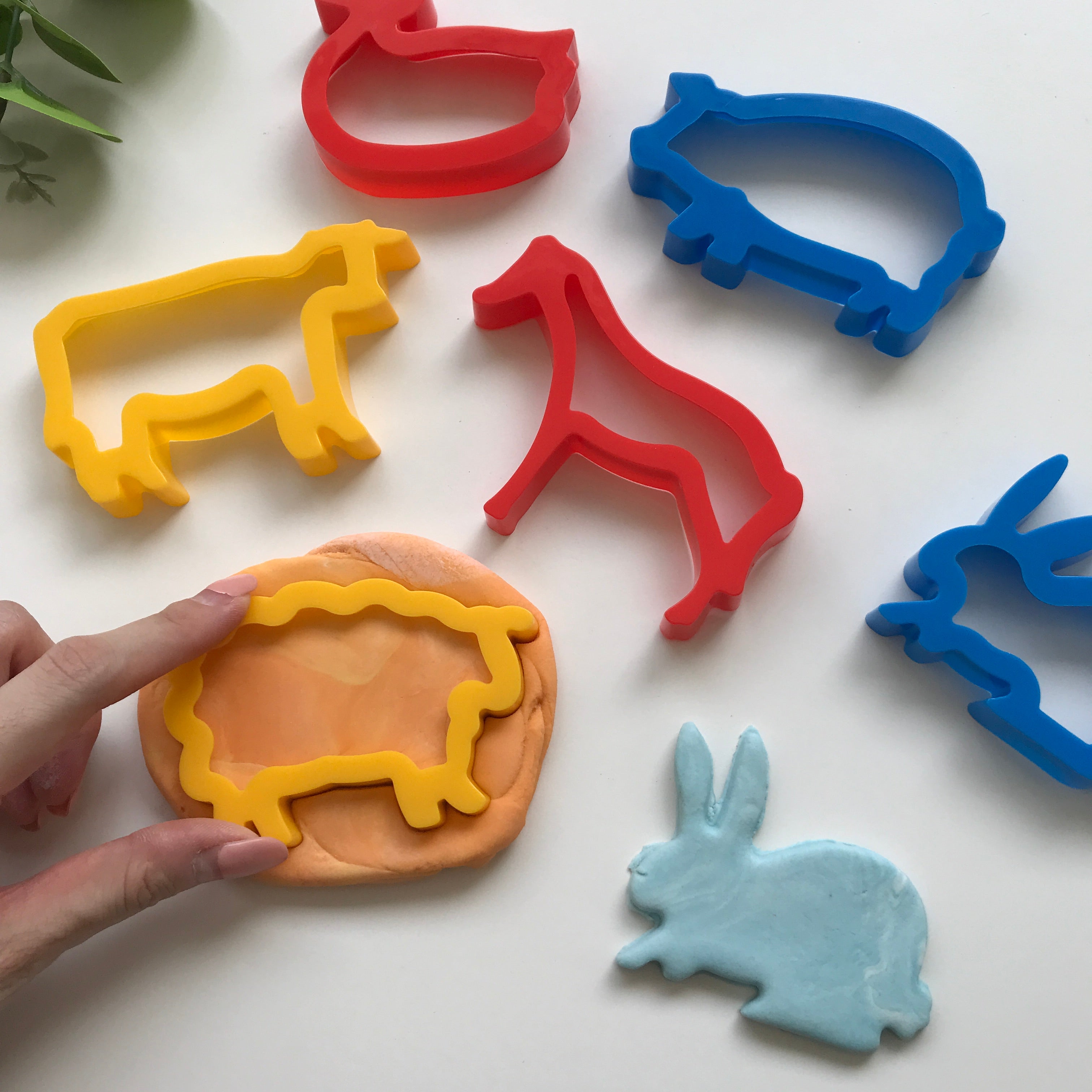 A set of colorful plastic playdough cutters in the shapes of various farm animals, including a horse, cow, sheep, duck, pig, and rabbit, displayed on a white background with a person's hand pressing a dough figure of a rabbit.