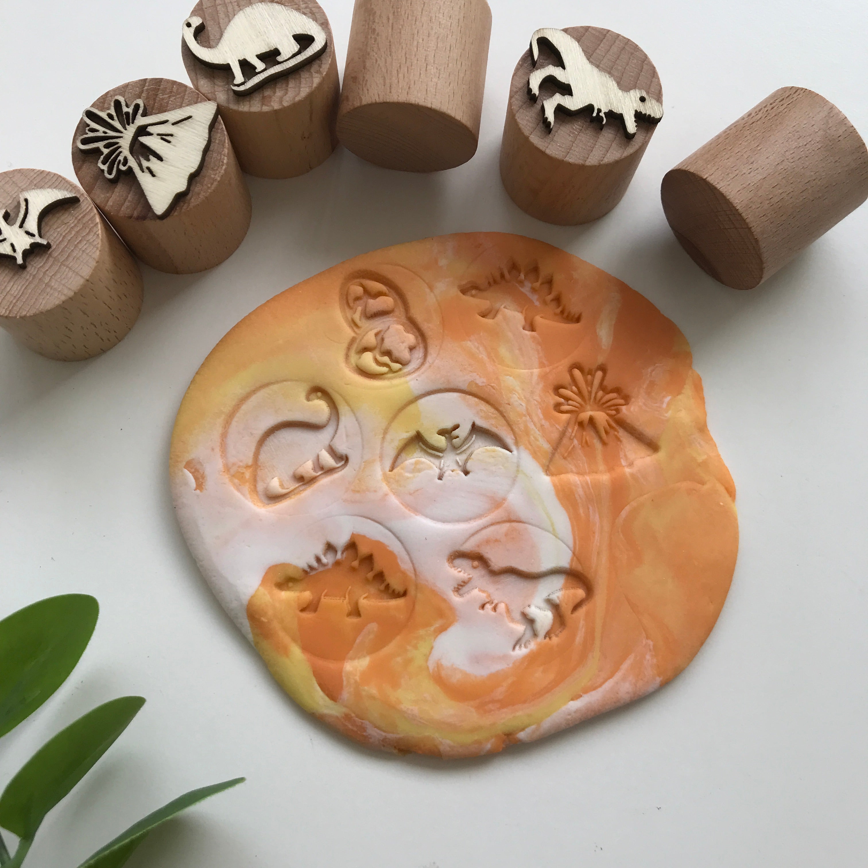 Set of 6 wooden dinosaur themed wooden stampers alongside their respective imprints on a marbled orange and white playdough