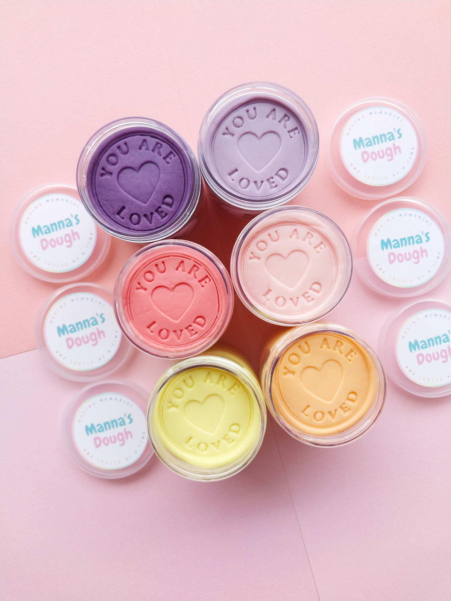 A group of Ssix tubs of playdough layed out flat, with their covers opened. The playdough colors are Purple, Lavender, Dark Pink, Light Pink, Light yellow, Pastel Orange. Playdoughs are stamped with the phrase 'You are Loved"