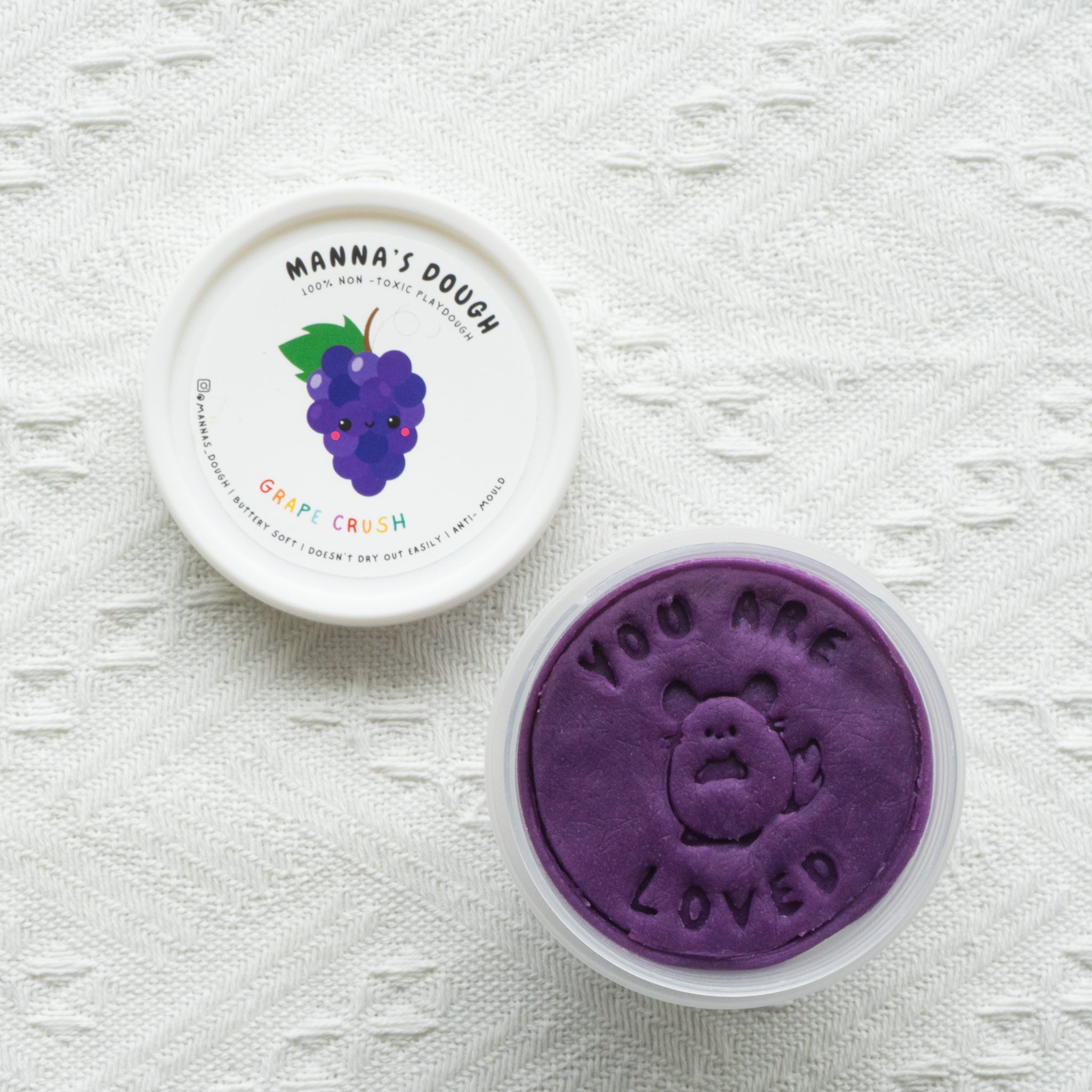 A container of purple playdough with a lid featuring a grape design, and the text 'You are loved' on the playdough surface.