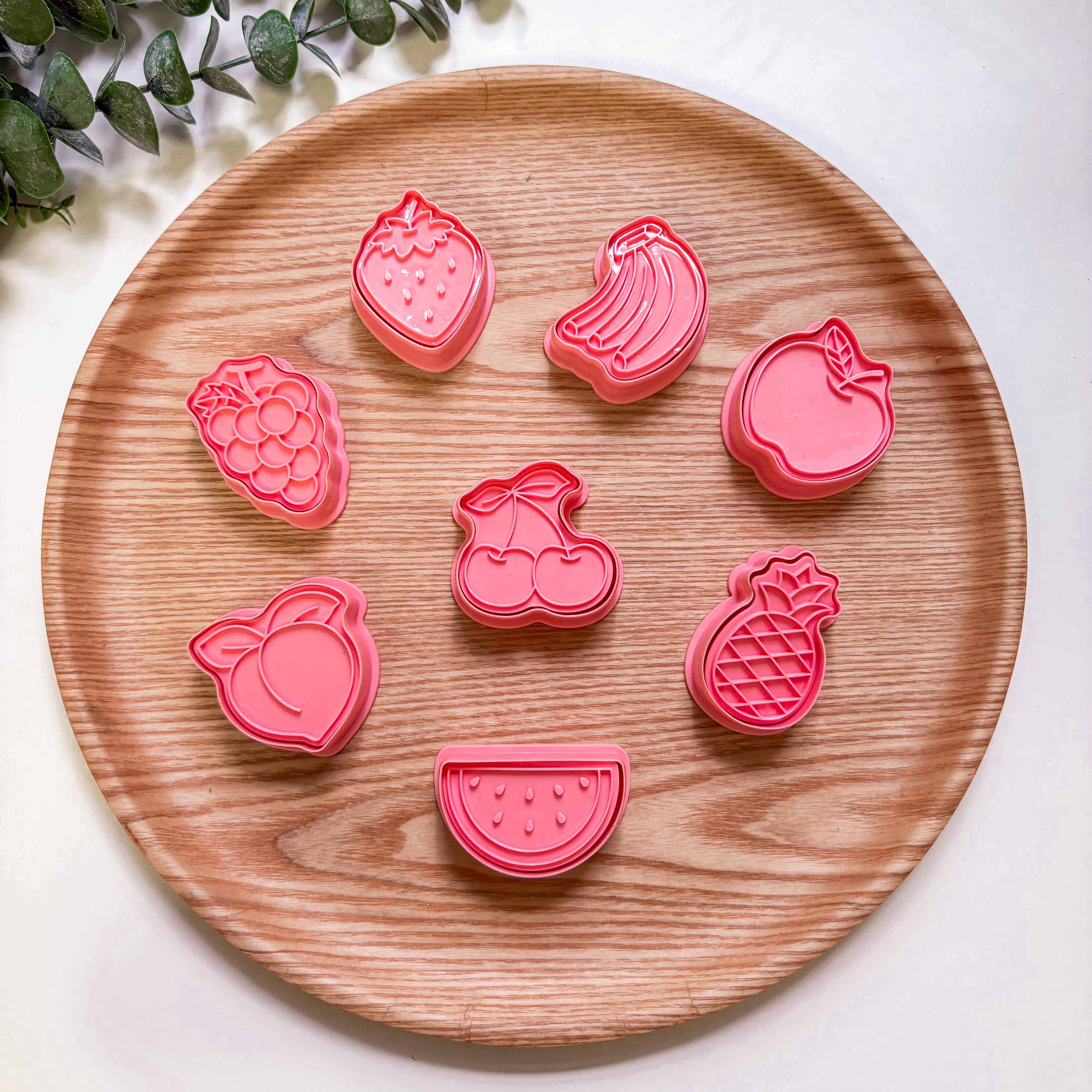 Set of pink fruit-shaped cookie cutters on a wooden plate with a white background.