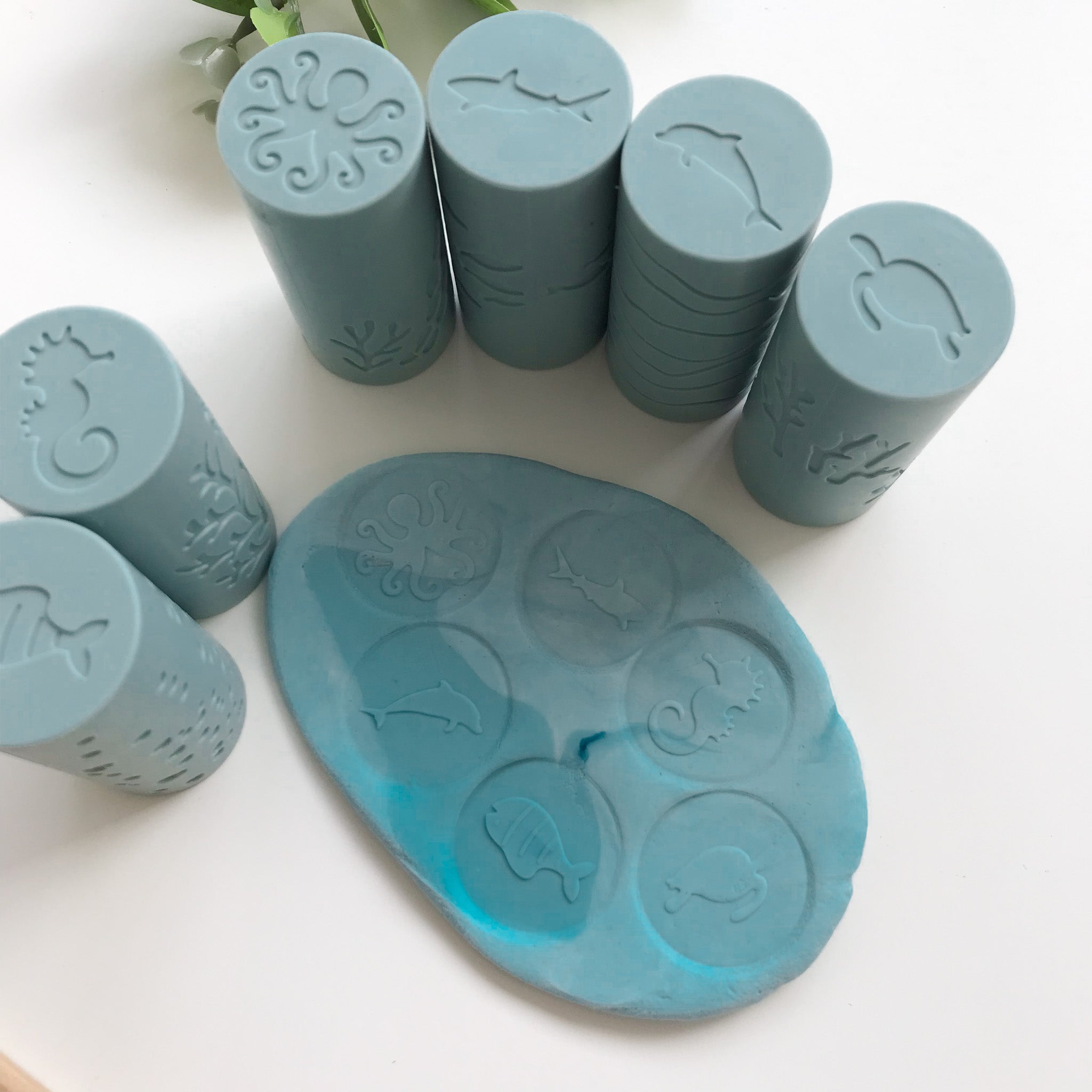 A set of 6 silicone playdough rollers and their respective imprints on a flat piece of blue playdough. The imprints formed are an octopus, shark, dolphin, seahorse, fish and turtle