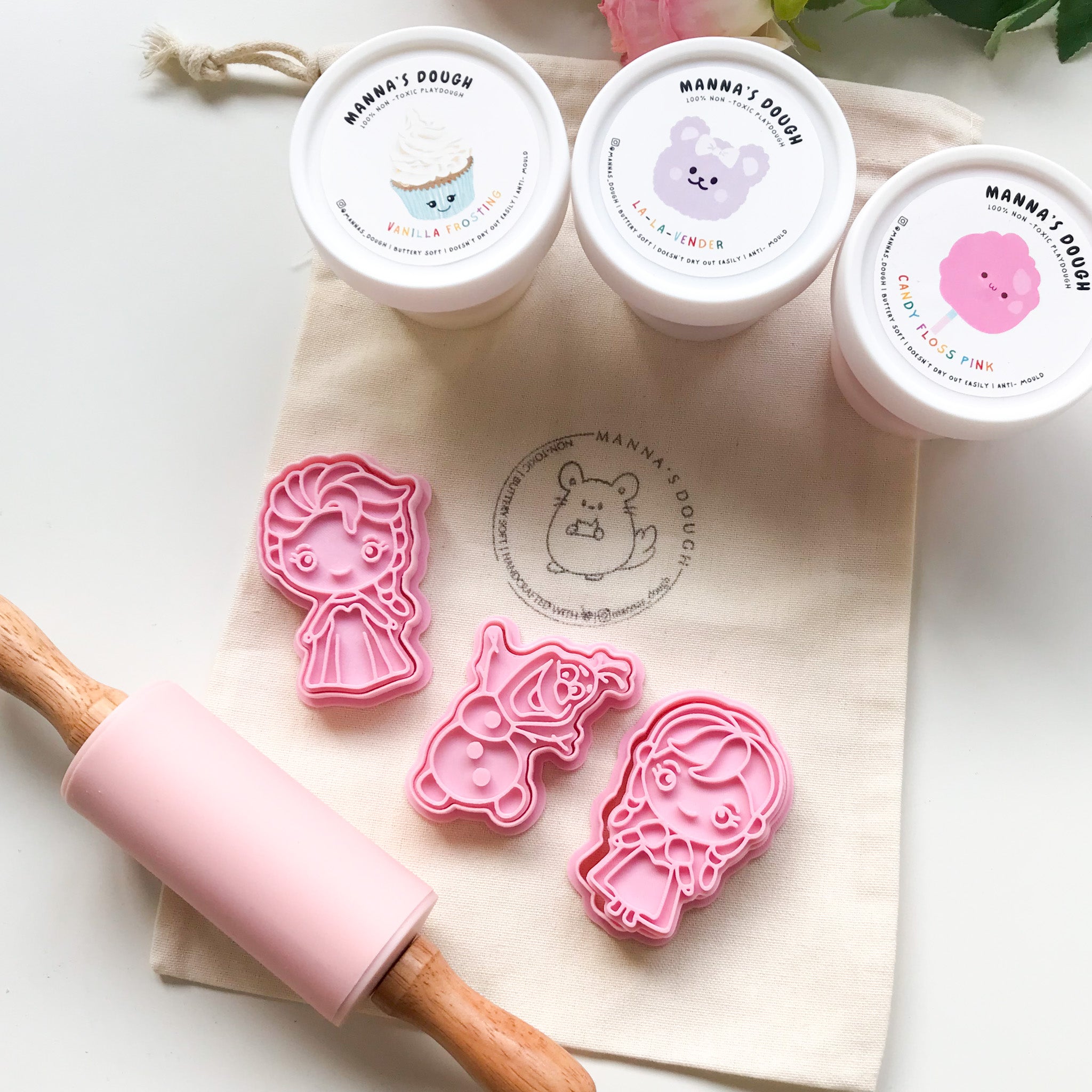Elsa playdough kit featuring 3 tubs of playdough with cute cartoons on the lid, a set of Frozen inspired stampers (Elsa, Olaf and Anna cutters) and a pink silicone playdough roller, all set against a canvas bag with the Mannas Dough Logo