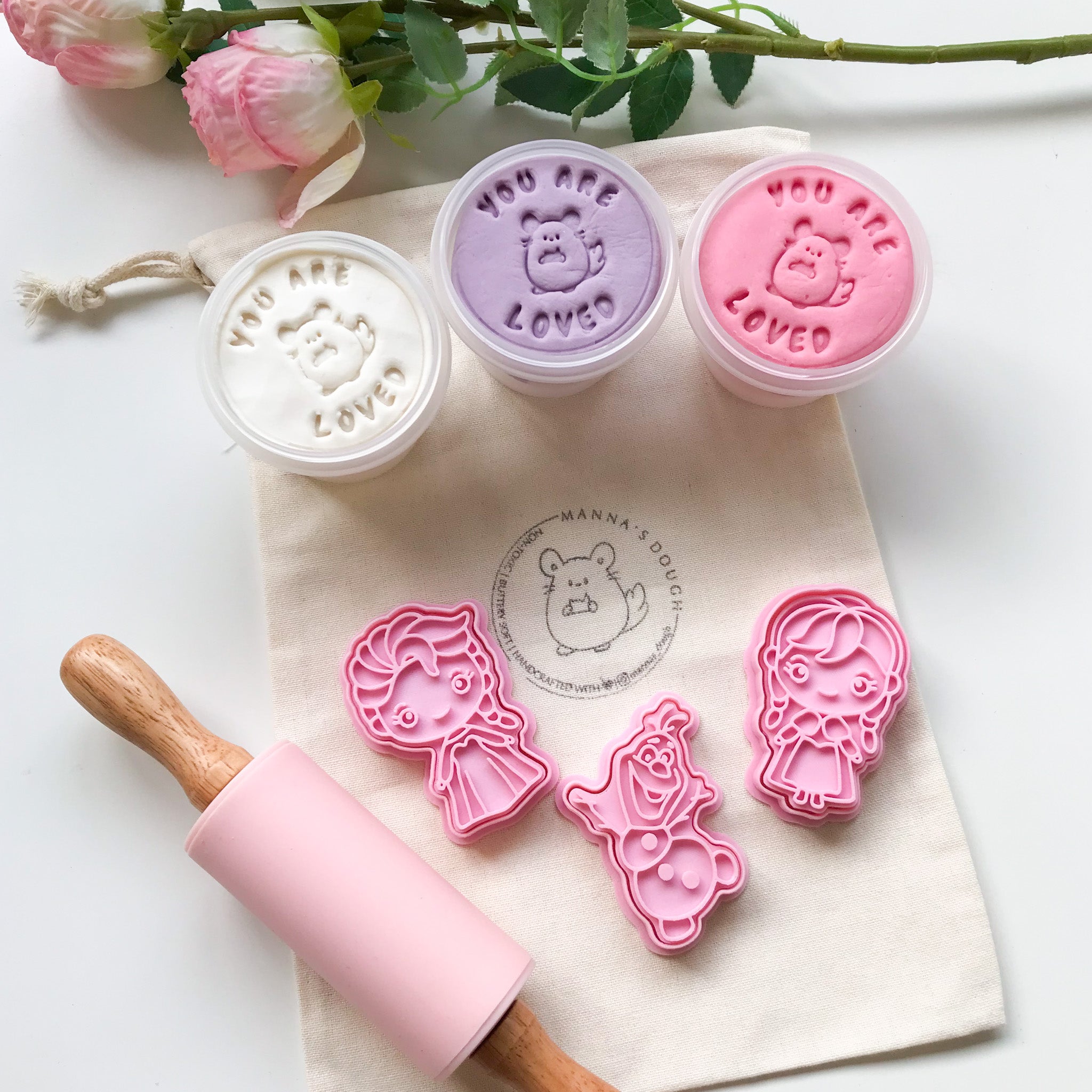 Elsa playdough set that features 3 tubs of playdough (white color, lilac color, pastel pink color), a set of Frozen themed stampers (Elsa, Olaf and Anna) and a pastel pink silicone roller. These items are set against a canvas bag with the Mannas Dough logo stamped on it