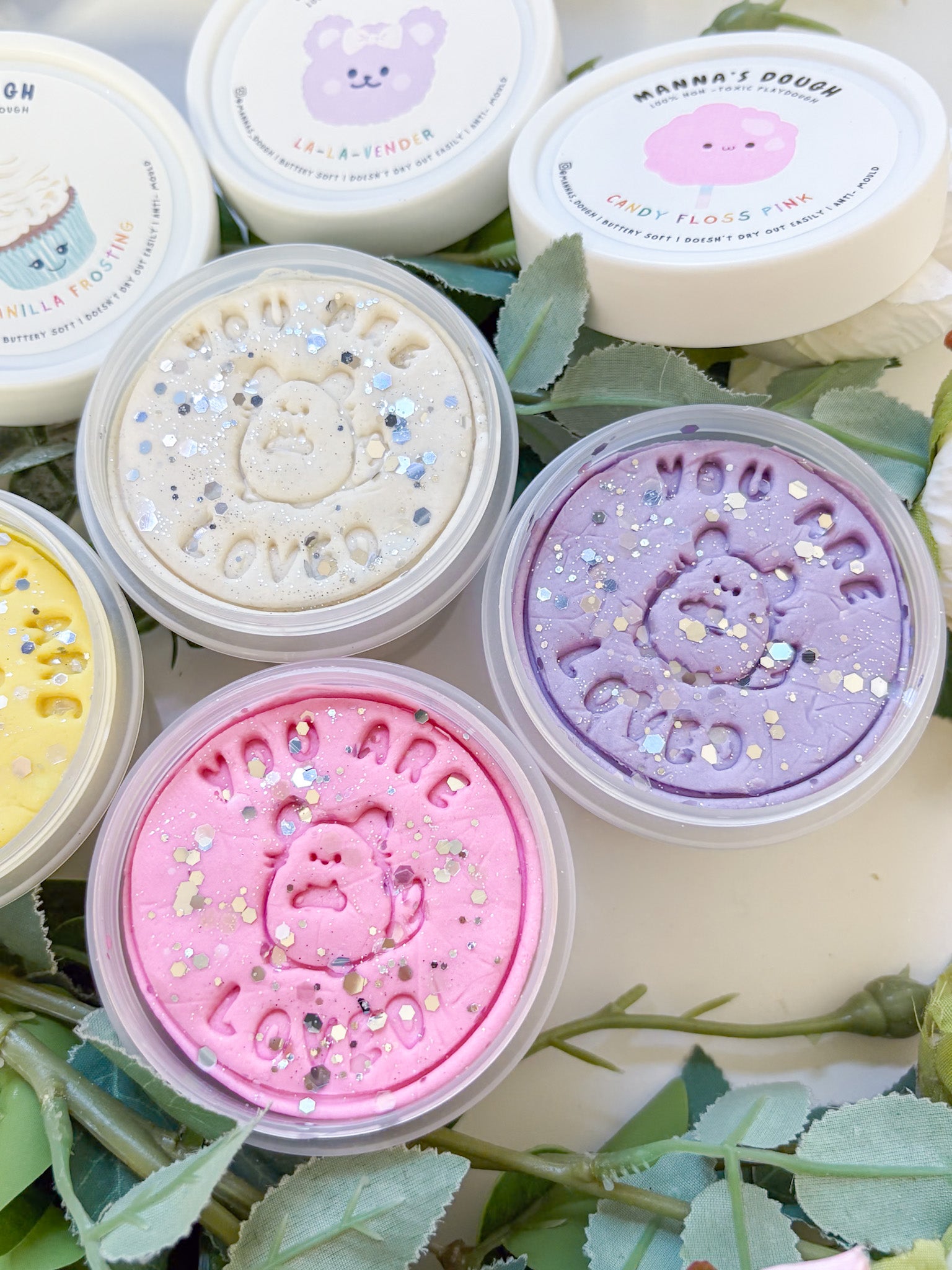 Glitter playdoughs in the colors lilac, white, yellow and pastel pink. Set against a lush green backdrop