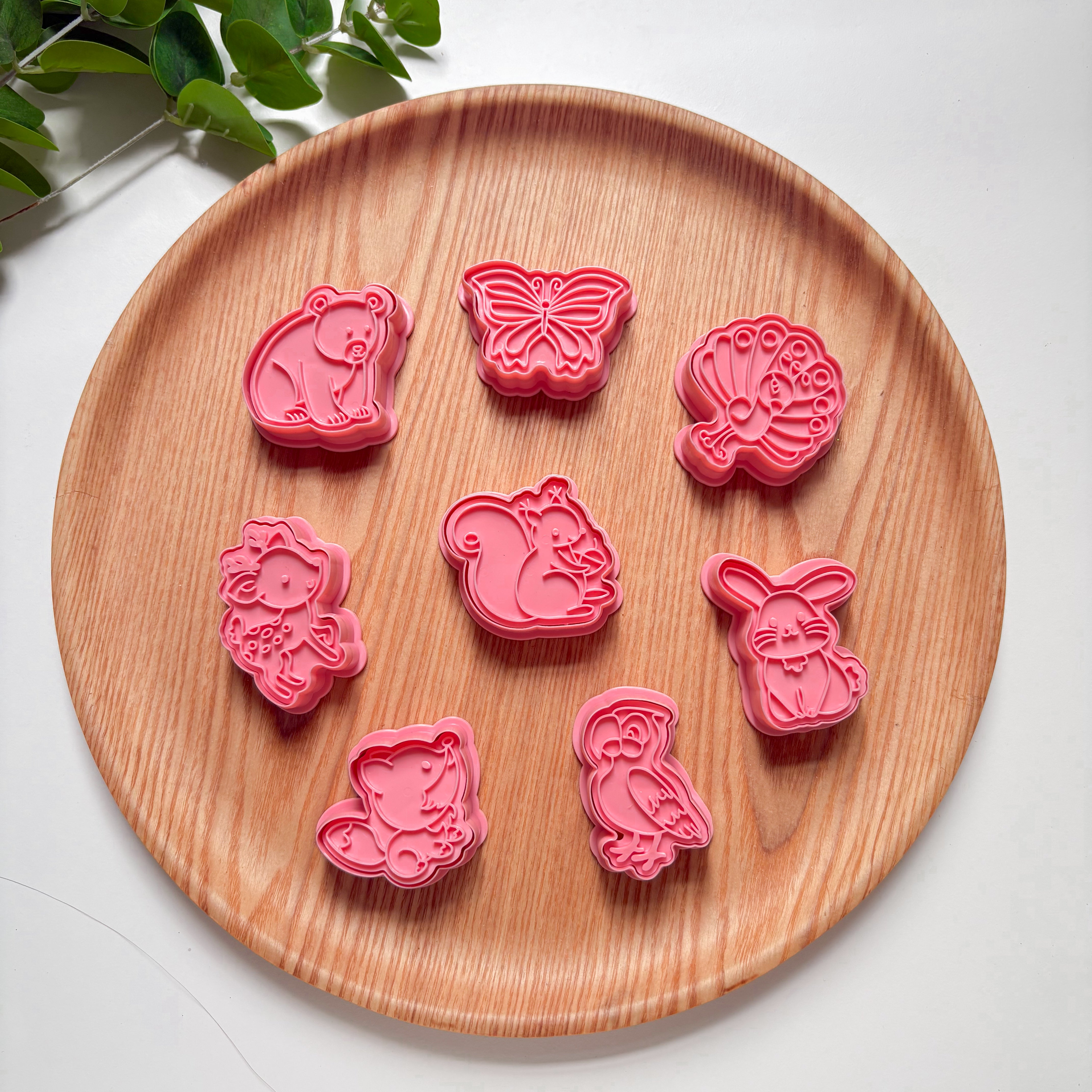 Set of pink animal-shaped playdough cutters on a wooden plate with green leaves in the background.