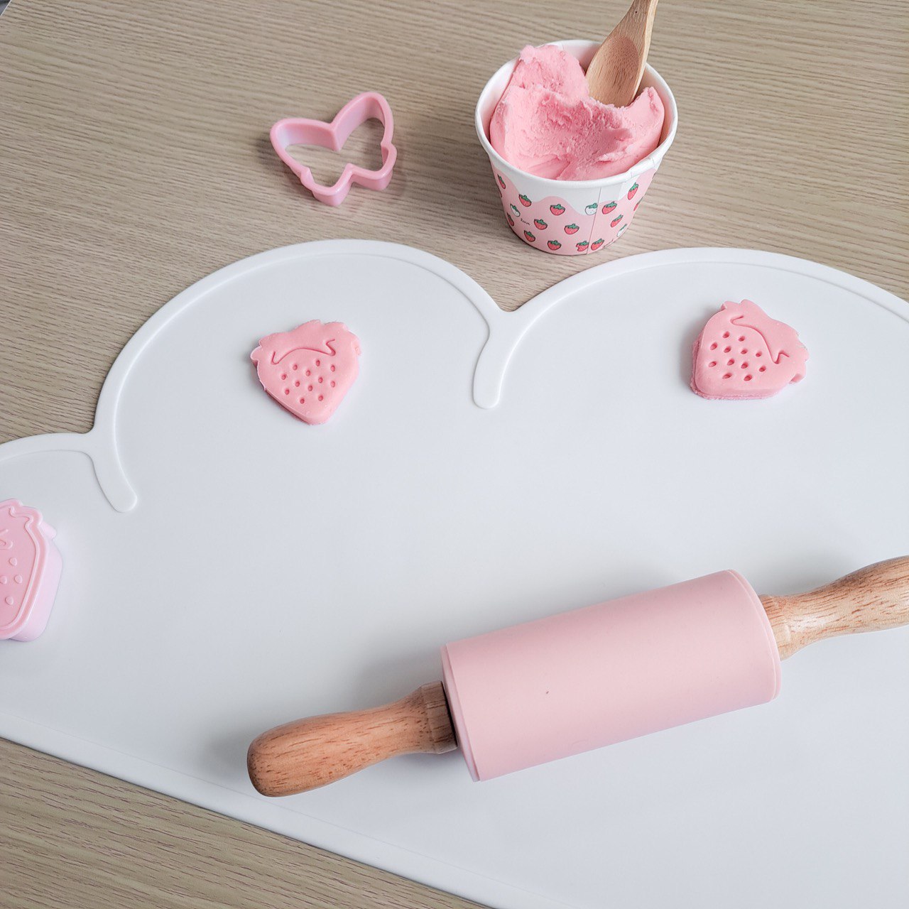 White silicone playdough mat, a pink roller and a tub of pastel pink playdough along with two paydough creations of strawberries