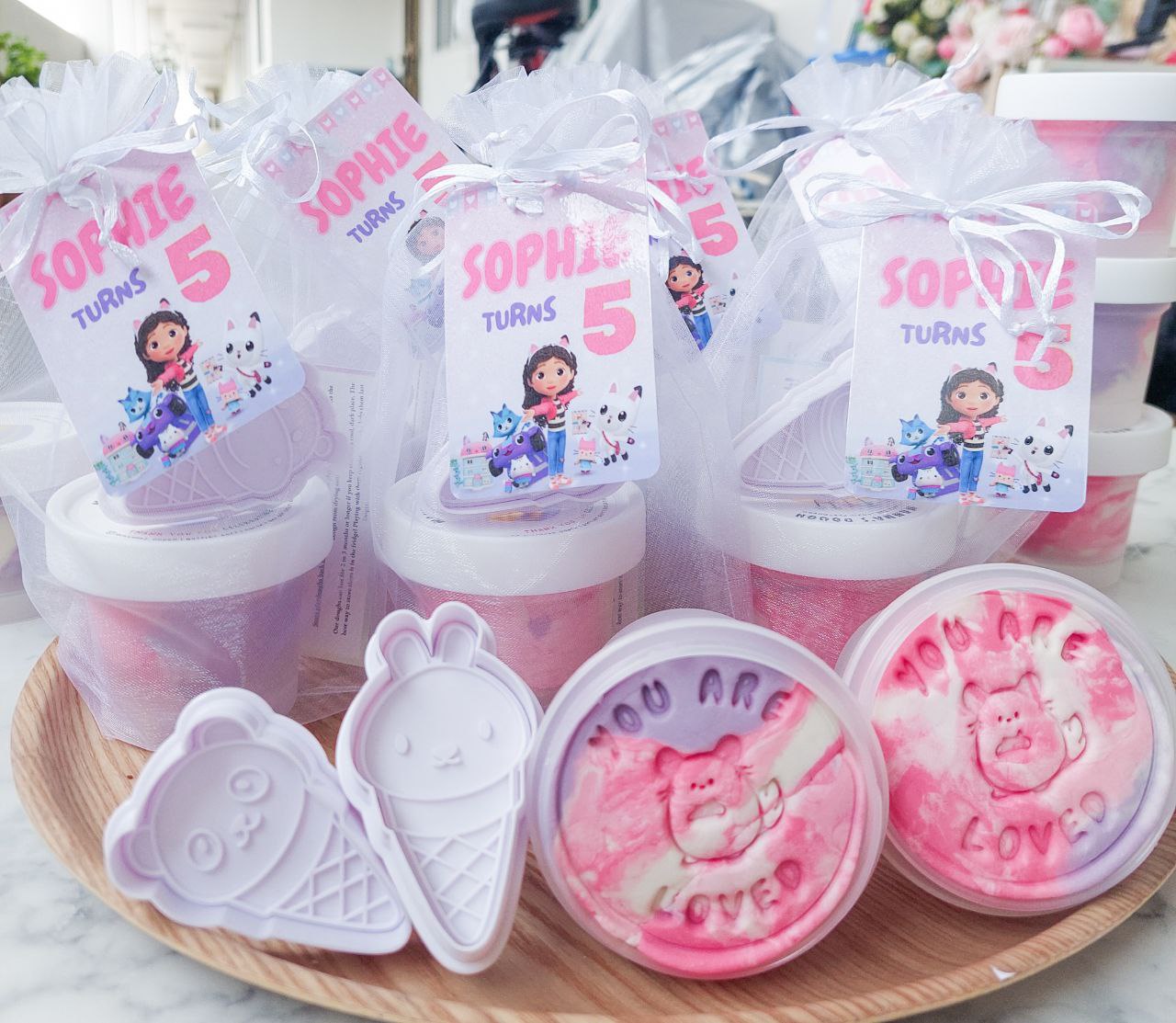 Party packs with marbled playdough design and themed tags displayed on a wooden tray, with a 'Gabby dollhouse' character design on the tags.