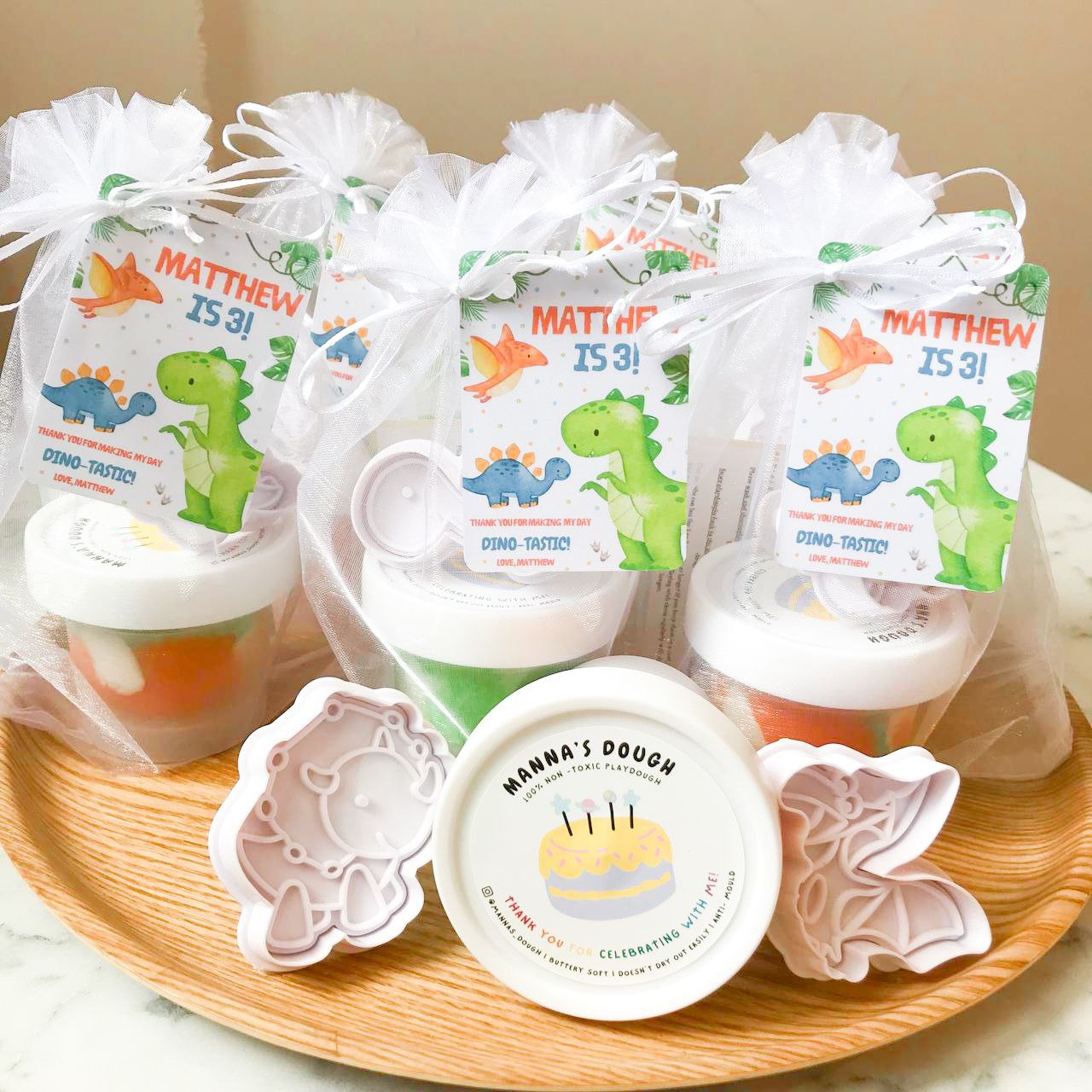 A set of dinosaur themed playdough party packs. Each pack featured a tub of marbled green, orange and white playduough alongside a dinosaur cutter