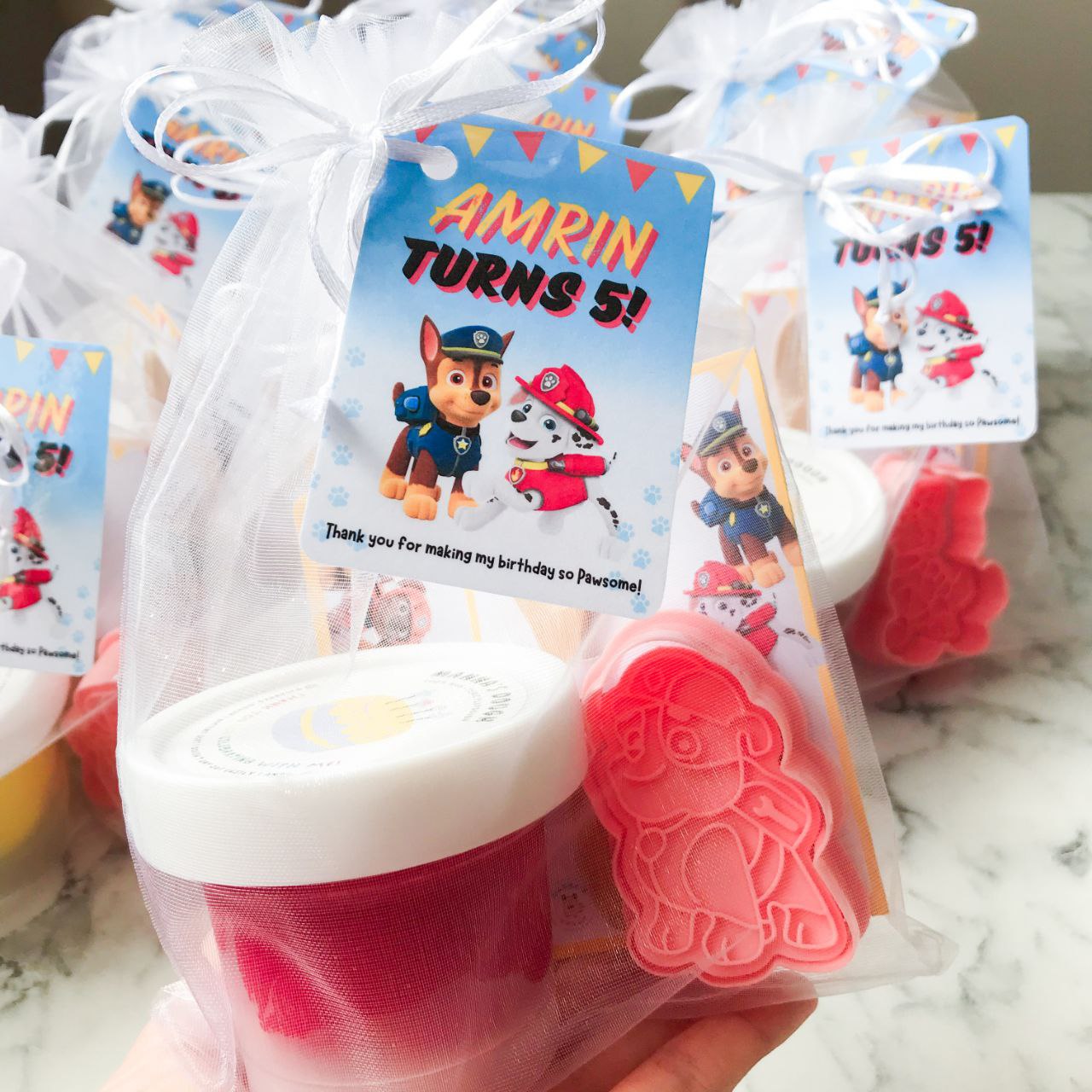 Packaged playdough party packs with Paw Patrol themed decorations, including character cutters and a themed gift tag.