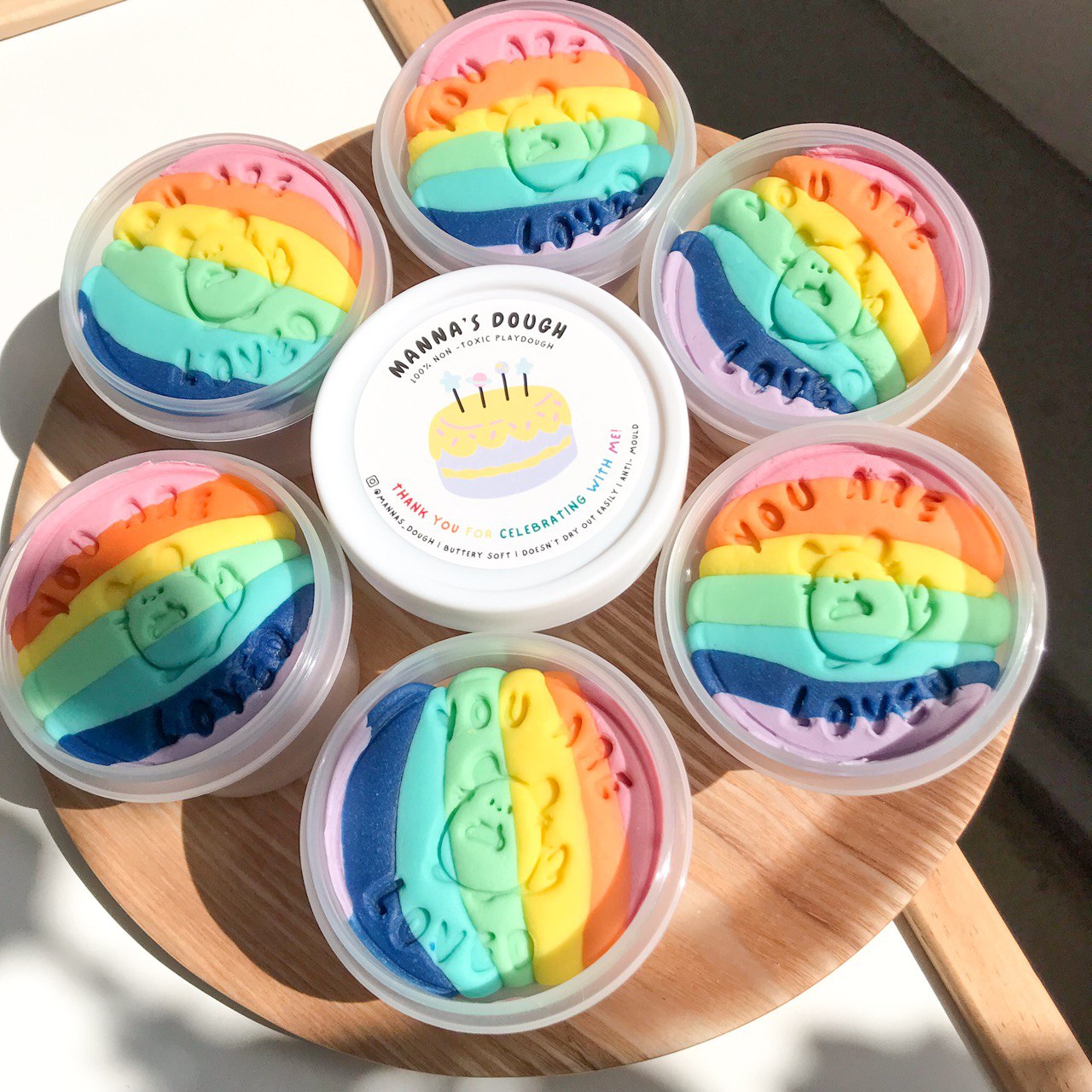 six tubs of multicolored non-toxic playdough arranged on a wooden surface, each with a rainbow pattern and a label in the center that features a birthday cake