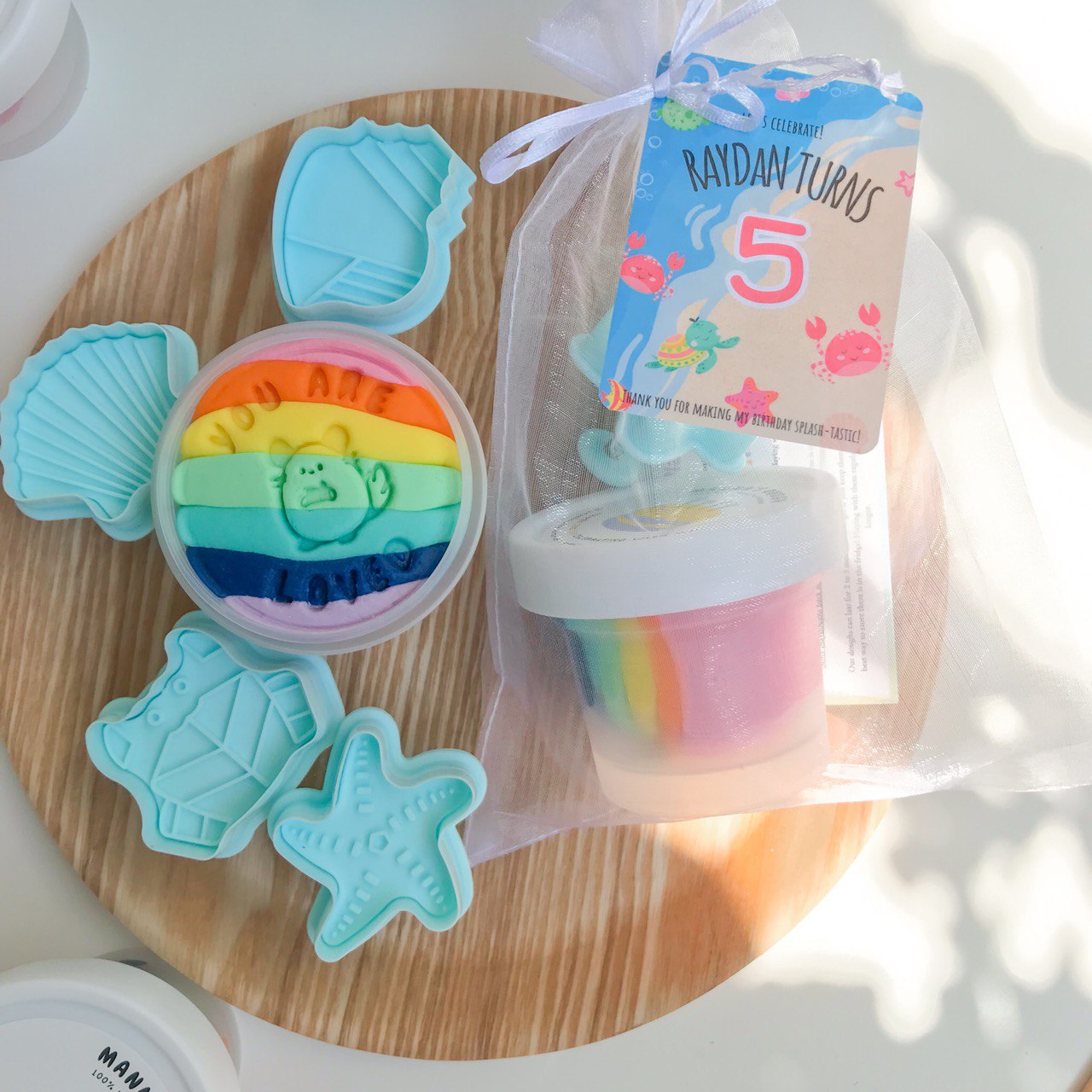 Flatlay photo of rainbow party pack. Contains 1 tub of rainbow playdough and a set of 4 ocean themed playdough stampers. There is also a party pack that contains one tub of playdough and a stamper packaged in a organza bag and a ocean gift tag