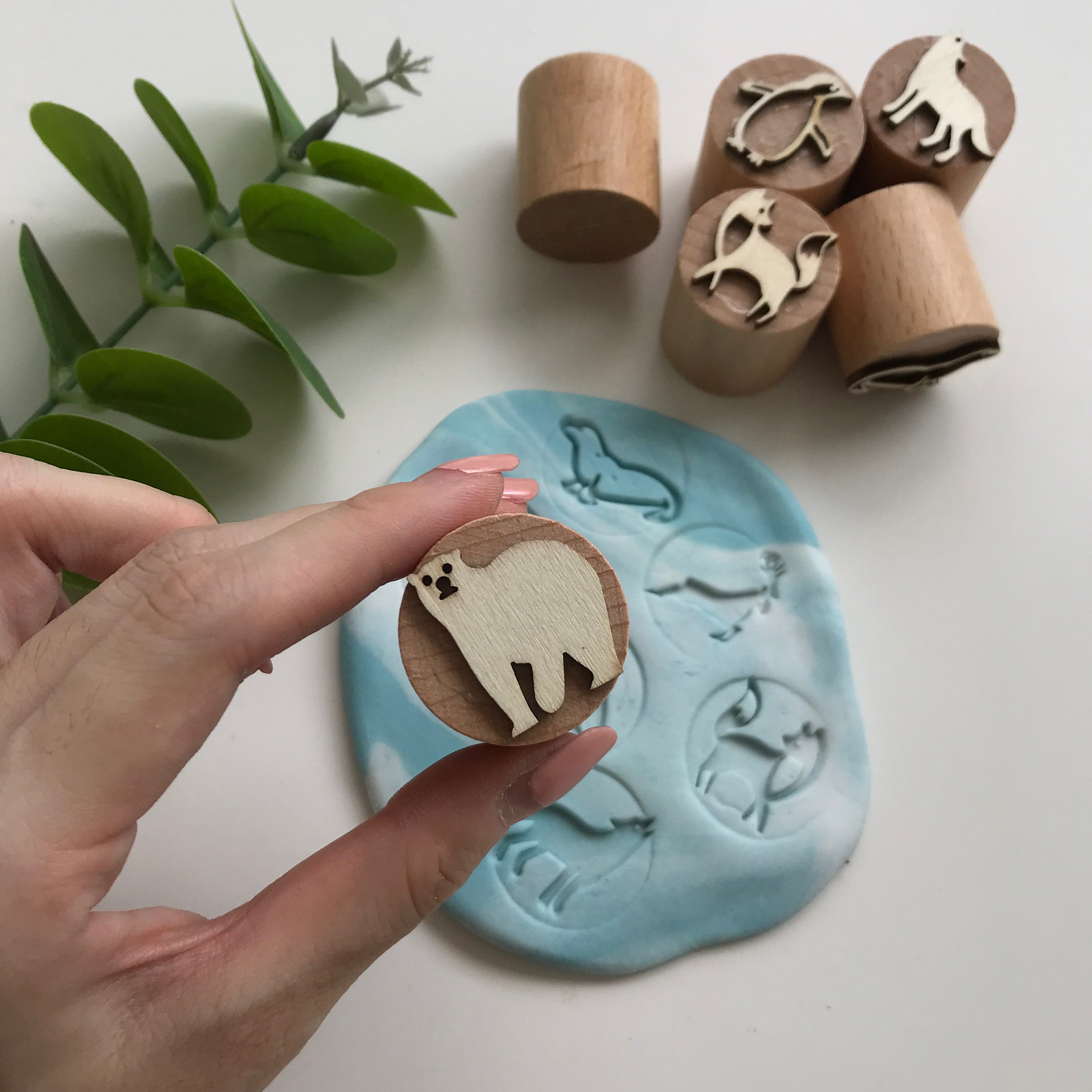 Hand holding a wooden playdough stamper with a bear design. In the background are 5 other wooden stampers as well as a marbled white and blue playdough circle