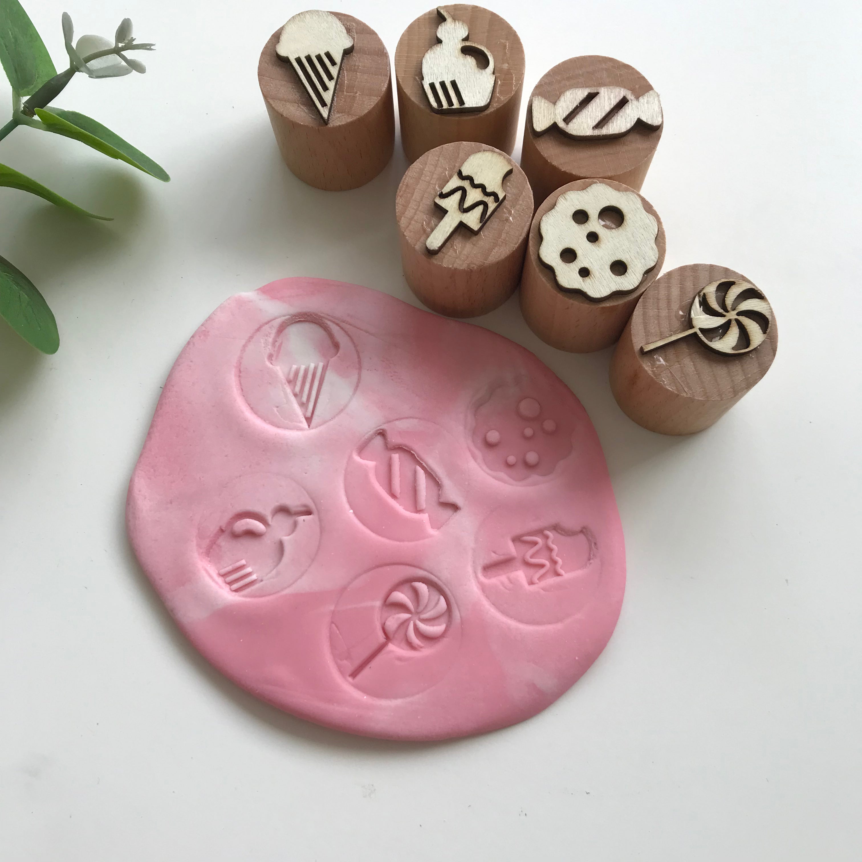 A set of six wooden stamps with shapes of ice cream, a cupcake, and other sweet items, alongside a pink piece of playdough featuring imprints of the playdough stamps