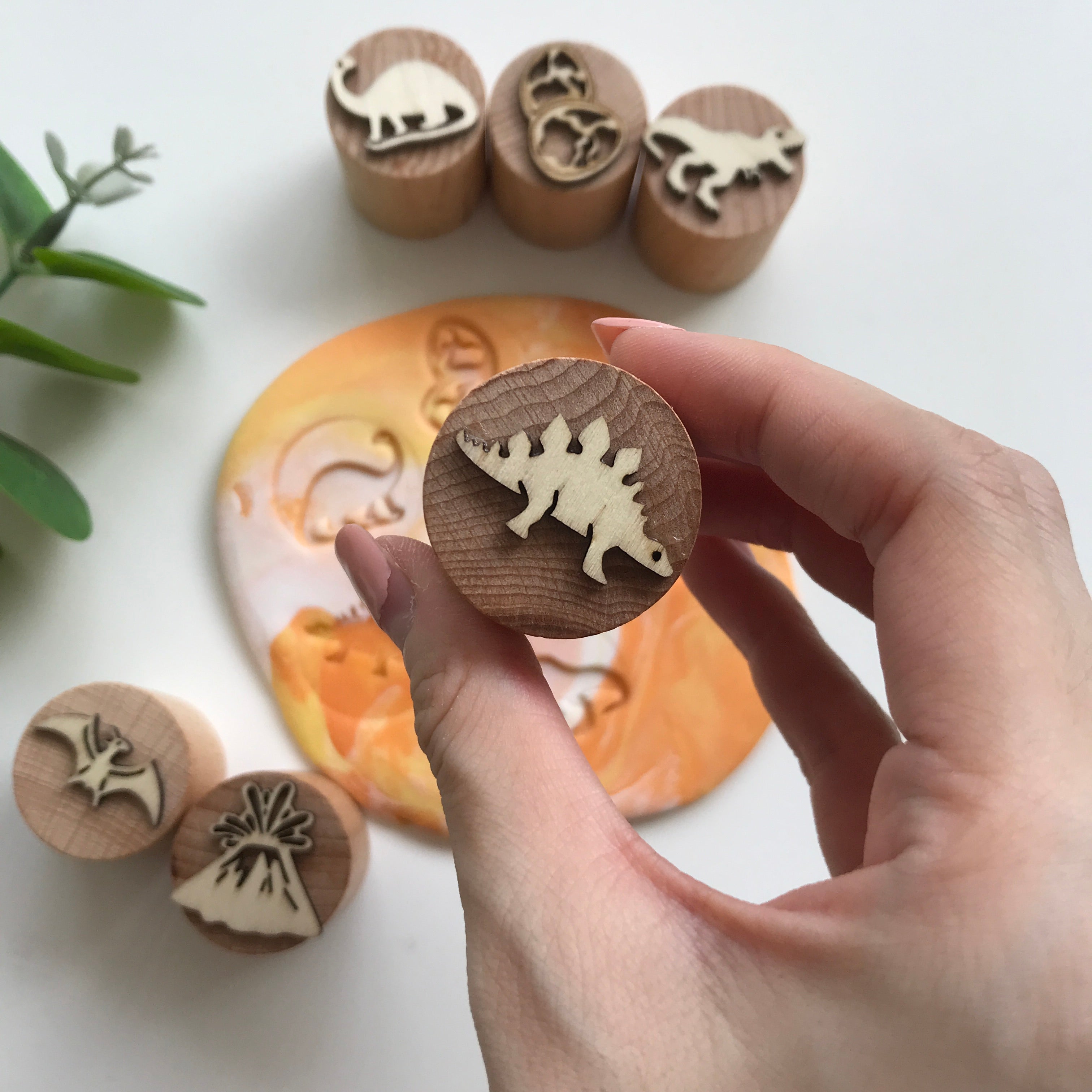 A hand holding up a stegosaurus wooden playdough stamp. In the background are other stamps with the following designs - pterosaur, volcano, t-rex, dinosaur eggs, Brachiosaurus
