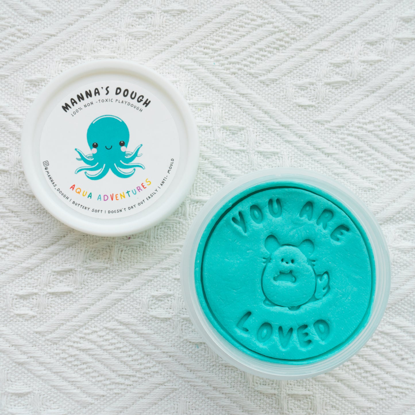 Bright turquoise playdough from Manna's Dough, labeled 'Aqua Adventures' with a cute octopus design on the lid. The playdough is stamped with 'YOU ARE LOVED' and an adorable animal character, perfect for kids' creative fun.