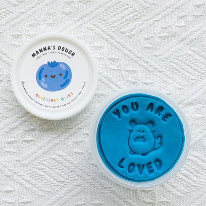 Deep blue playdough from Manna's Dough, labeled 'Blueberry Bliss' with a smiling blueberry design on the lid. The soft, non-toxic playdough is stamped with 'YOU ARE LOVED' and a sweet animal character, perfect for imaginative and safe play