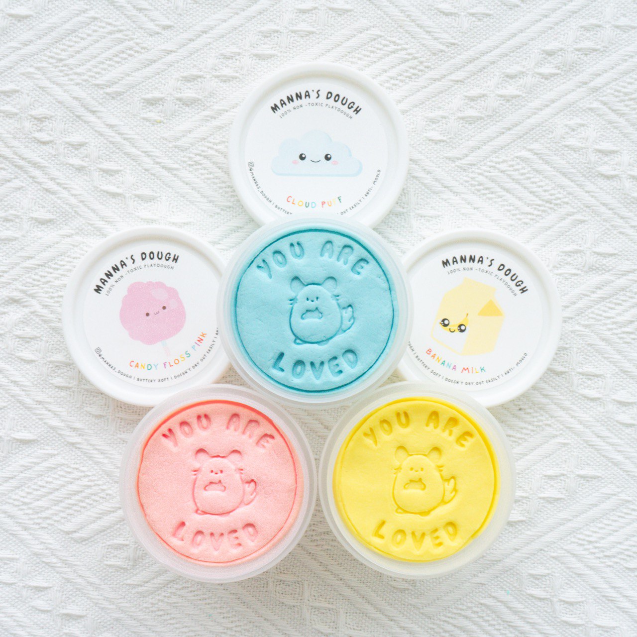 Flat lay of Manna's Dough playdough tubs arranged on a white textured background, featuring vibrant pastel colors like blue, pink, and yellow. Each tub lid has cute designs like a smiling cloud, cotton candy, and banana, while the playdough is stamped with 'YOU ARE LOVED' alongside an adorable character.