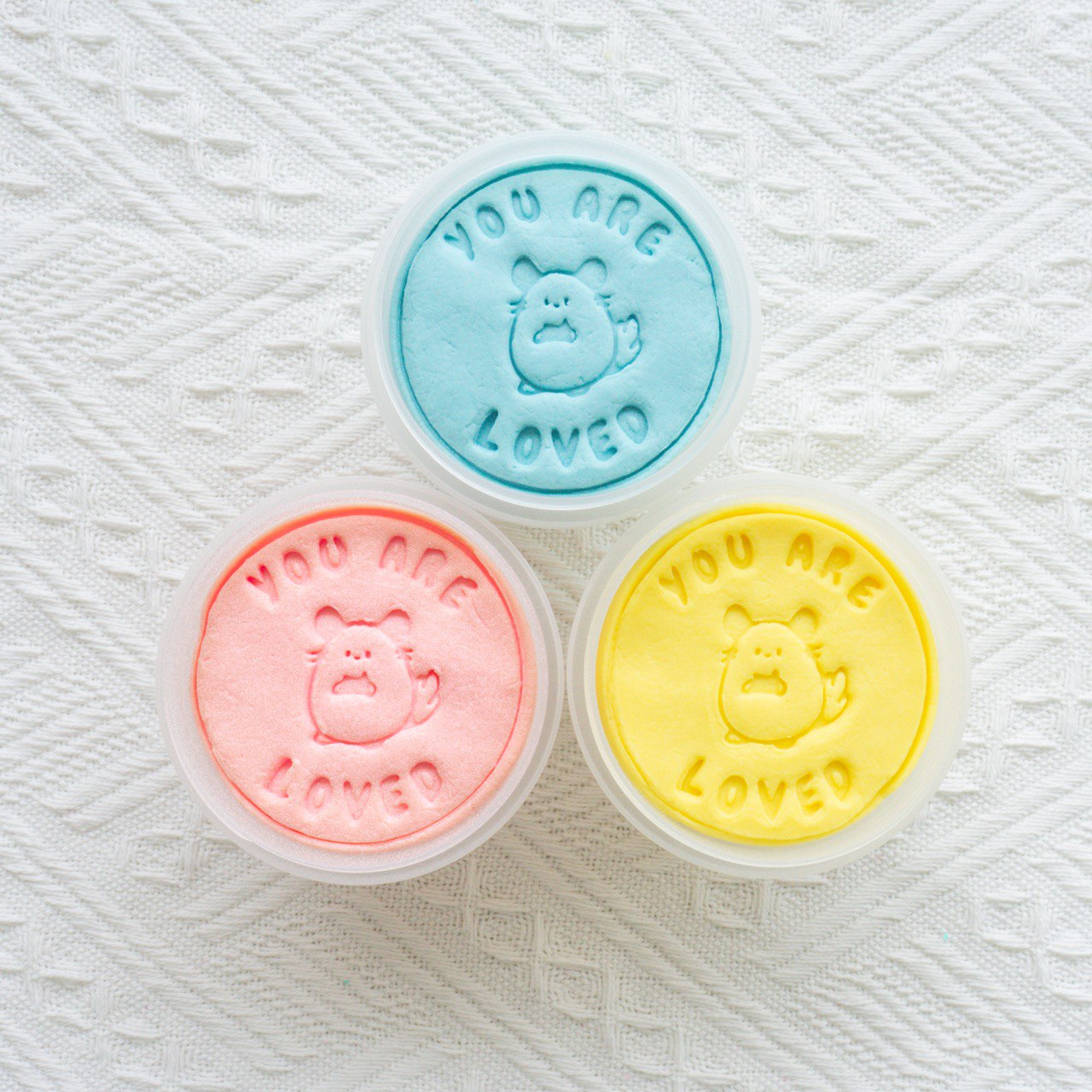 Close-up of three tubs of Manna's Dough playdough in pastel colors: blue, pink, and yellow. Each playdough piece is stamped with 'YOU ARE LOVED' and features a cute animal character, displayed on a white textured background