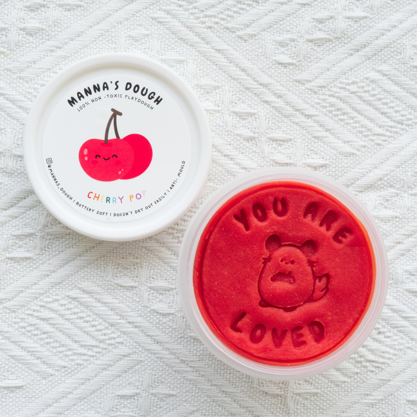 Vibrant red playdough from Manna's Dough, labeled 'Cherry Pop' with a cheerful cherry design on the lid. The soft, non-toxic playdough is stamped with 'YOU ARE LOVED' and a cute cherry character, ideal for safe and creative playtime.