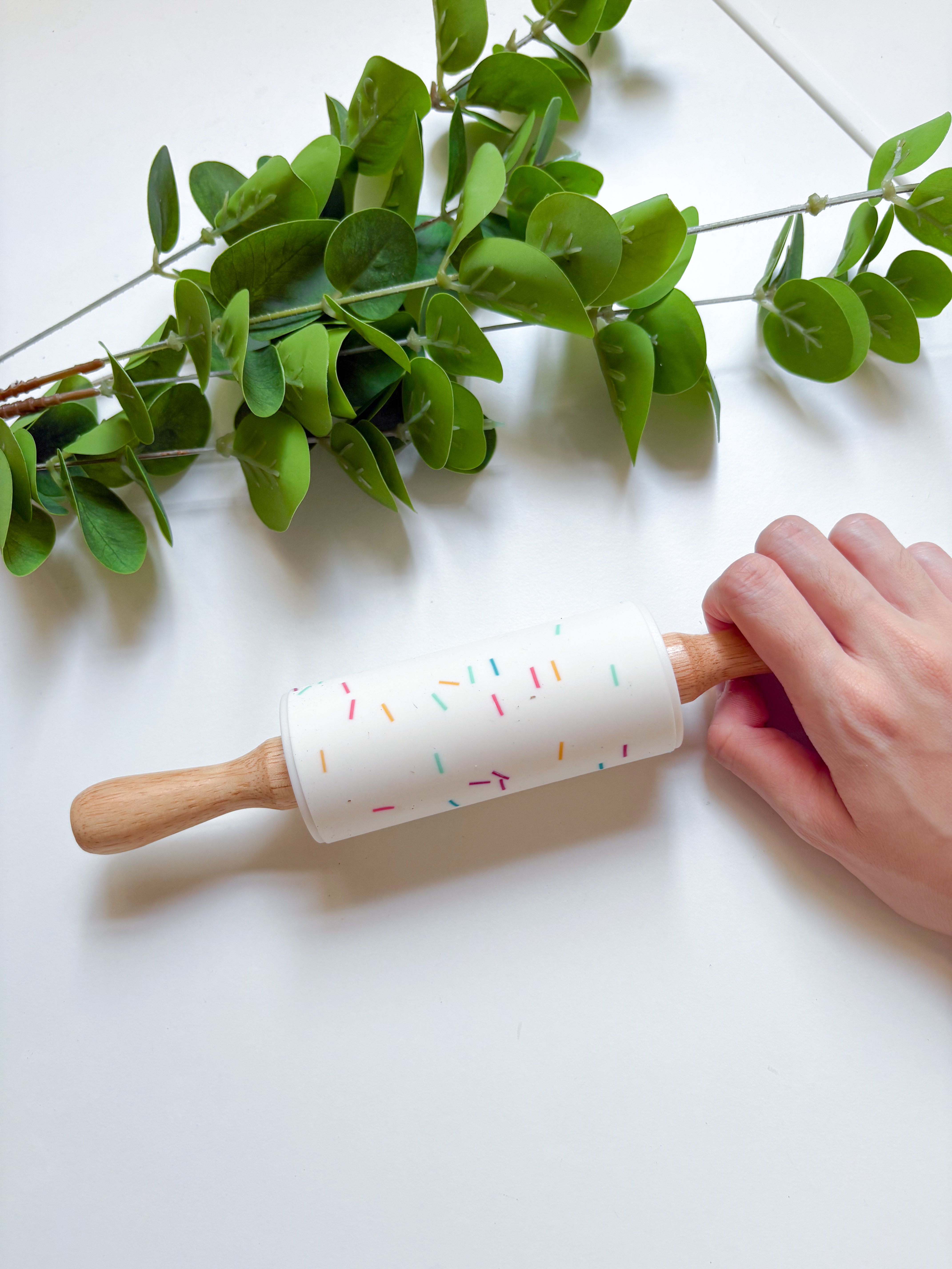 Hand holding a rolling pin with a decorated handle on a white surface with green leaves.