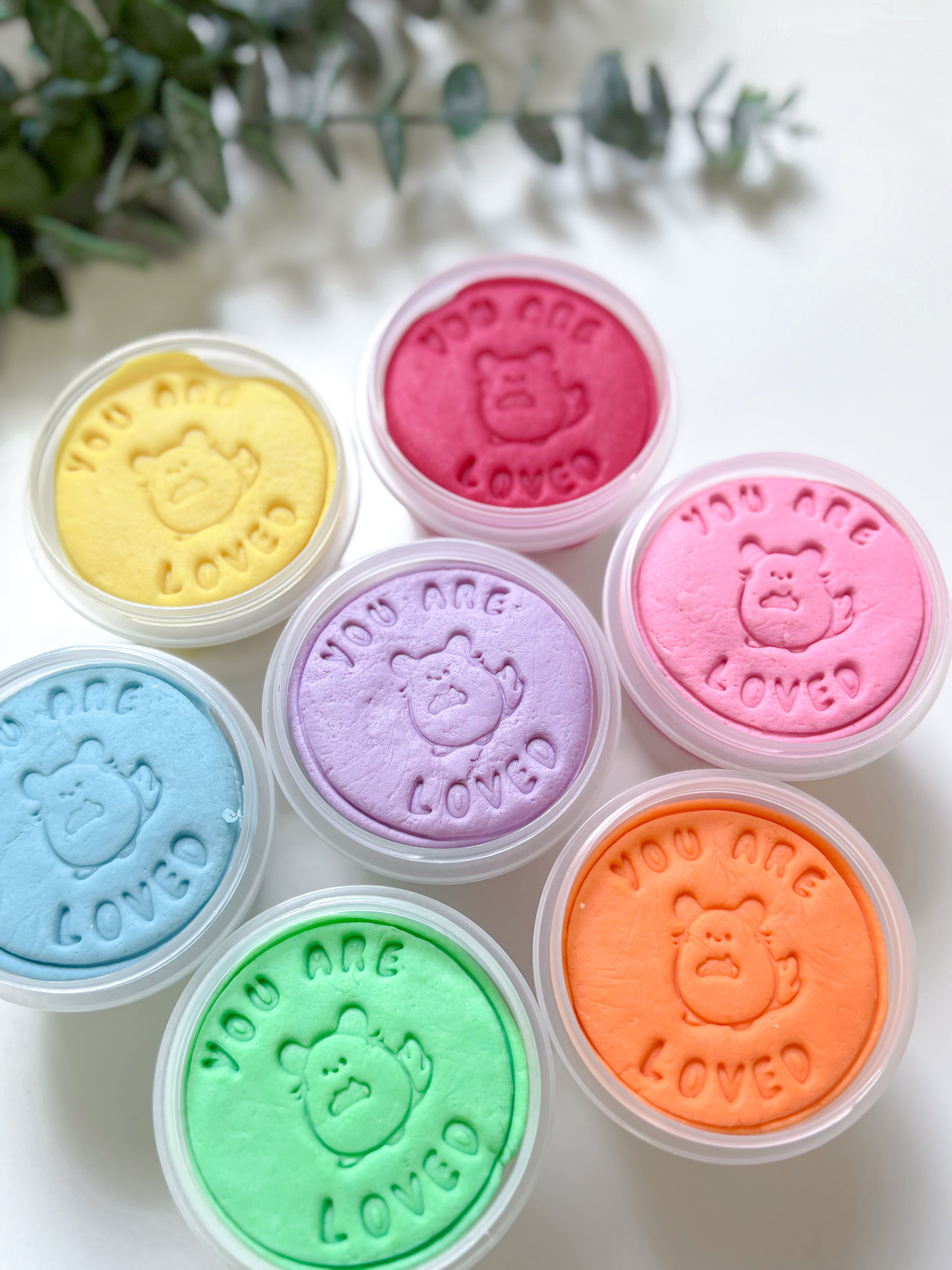 Set of colorful playdough tubs with 'You are loved' text and bear design on a light background.