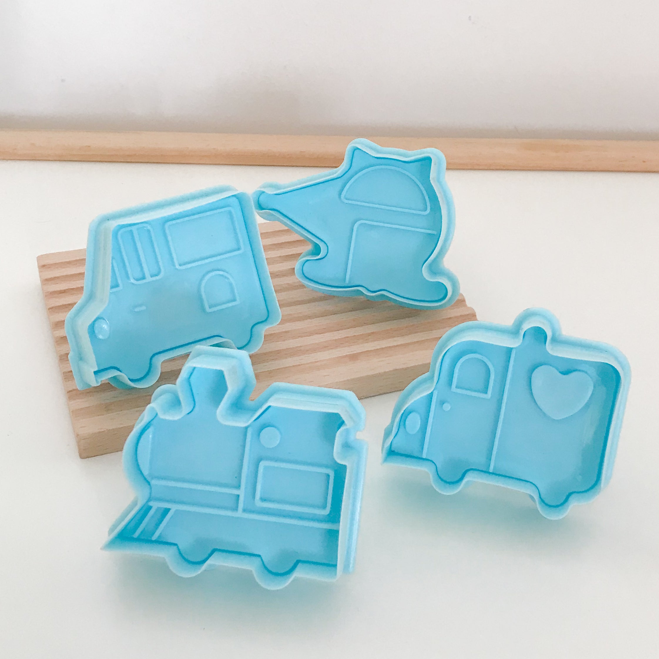 A set of four blue transportation-themed playdough stampers with shapes of a van, helicopter, train, and car, displayed on a wooden surface.