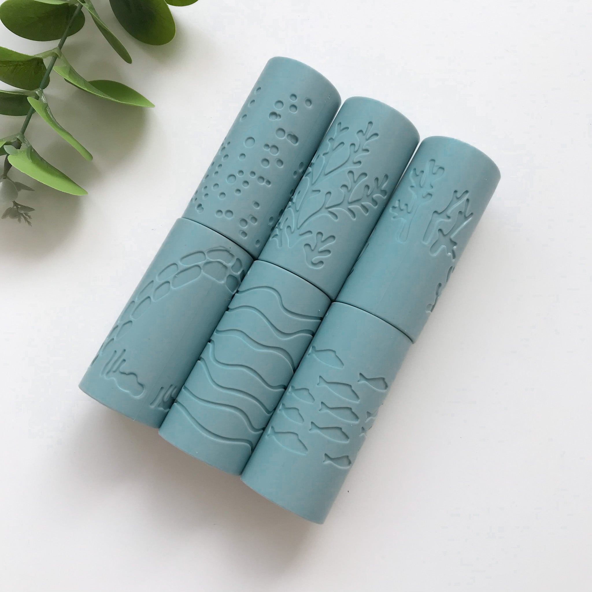 A set of blue silicone playdough rollers with ocean-themed designs, placed on a white surface, accompanied by a green plant 
