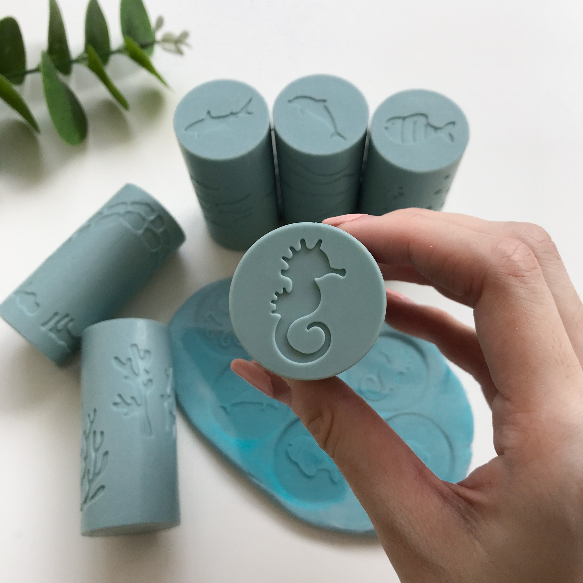 A hand holding up a silicone playdough roller that features a seahorse print. There are 5 other rollers in the background