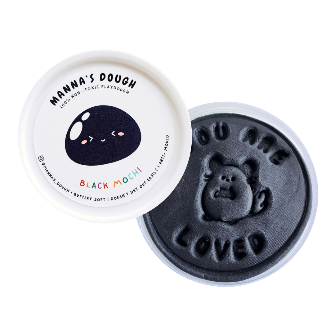 A container of Manna's Dough playdough with the lid off, showing the black mochi-textured playdough and the brand's logo with a cute character design.