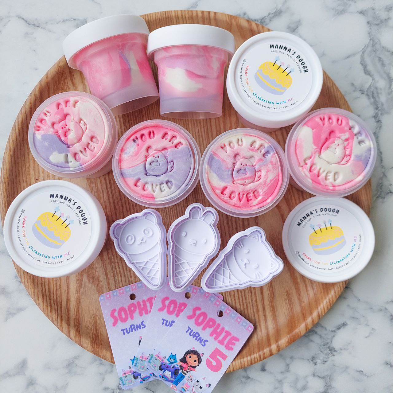 Party packs with marbled playdough design and themed tags displayed on a wooden tray, with a 'Gabby dollhouse' character design on the tags.