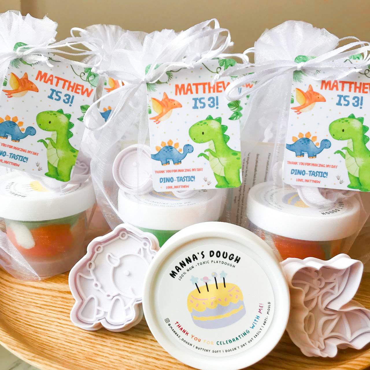 A set of dinosaur themed playdough party packs. Each pack featured a tub of marbled green, orange and white playduough alongside a dinosaur cutter