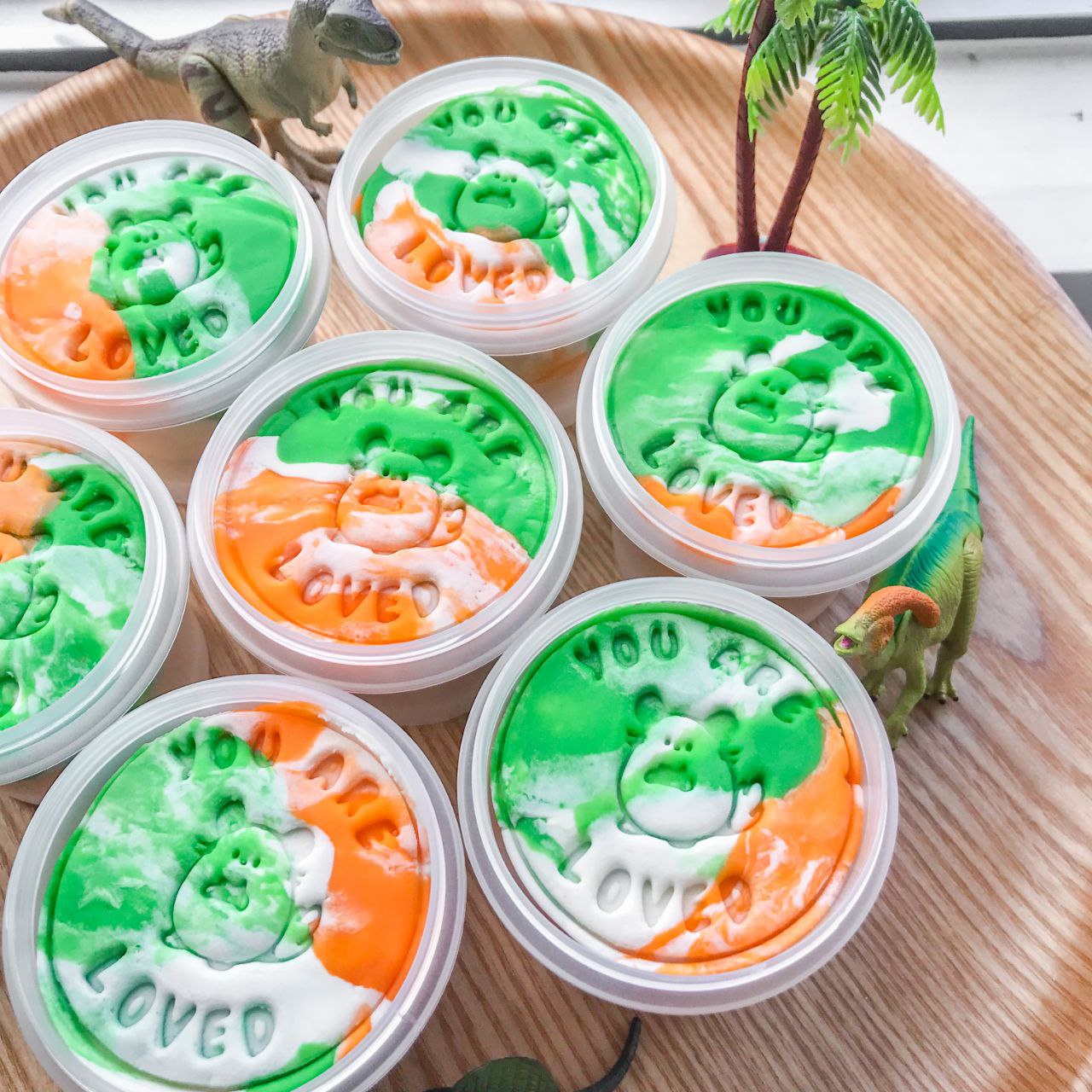 A set of dinosaur themed playdough party packs. Each pack featured a tub of marbled green, orange and white playduough alongside a dinosaur cutter