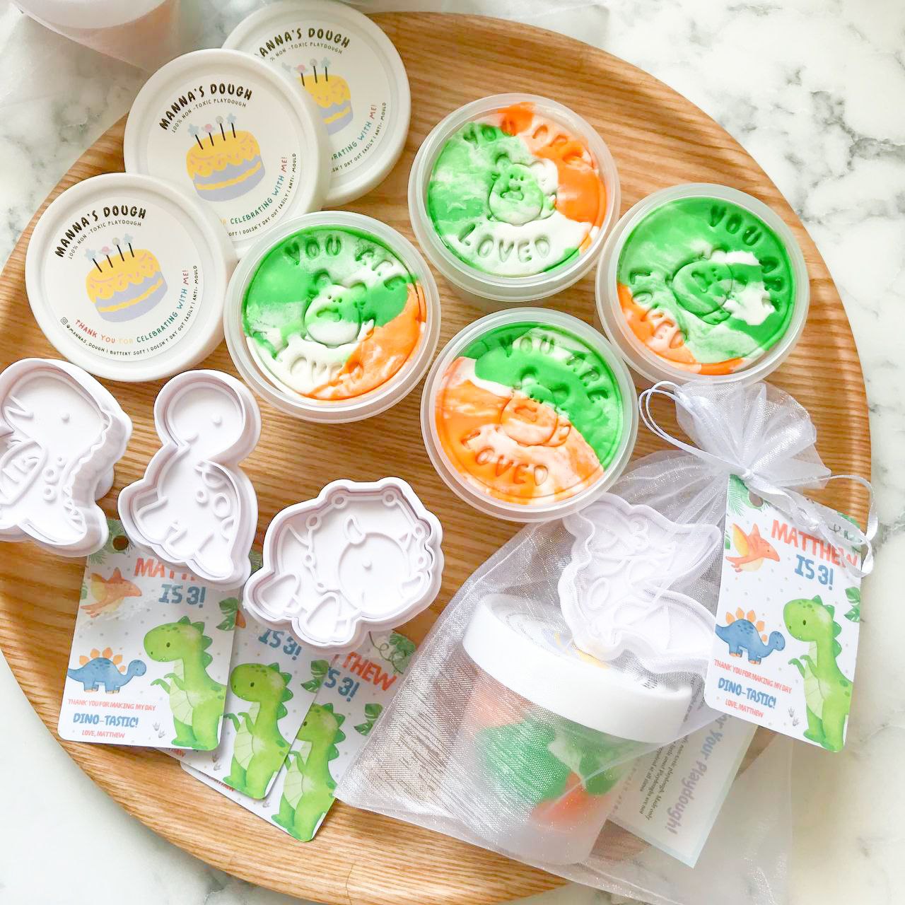 A set of dinosaur themed playdough party packs. Each pack featured a tub of marbled green, orange and white playduough alongside a dinosaur cutter