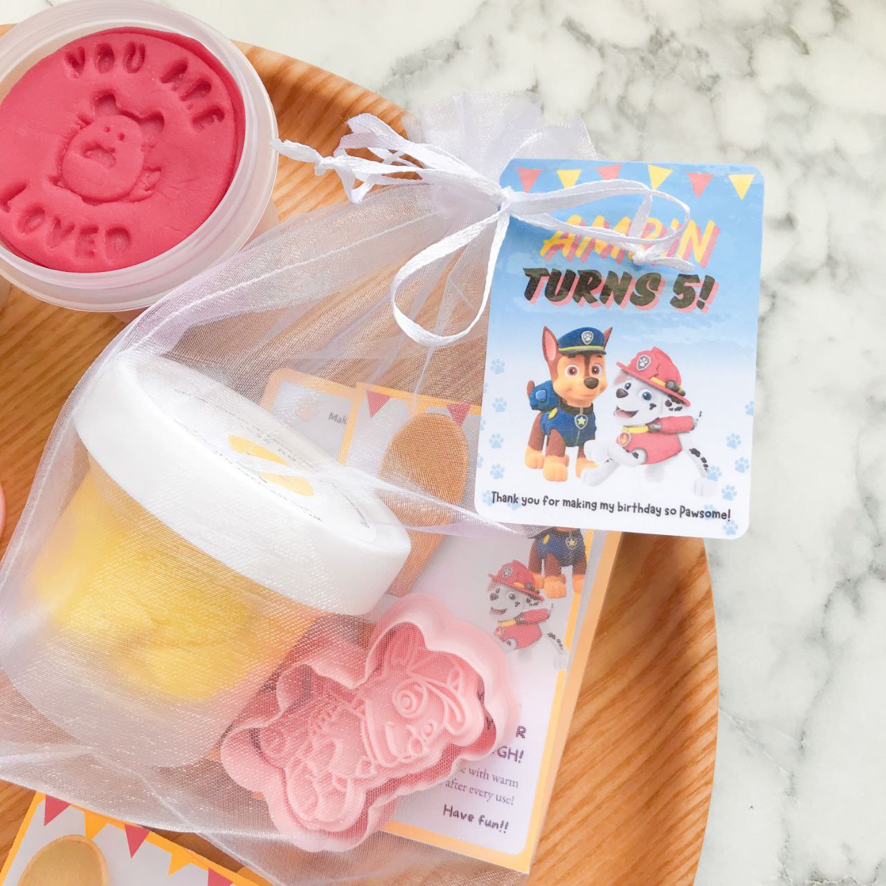 Packaged playdough party packs with Paw Patrol themed decorations, including character cutters and a themed gift tag.