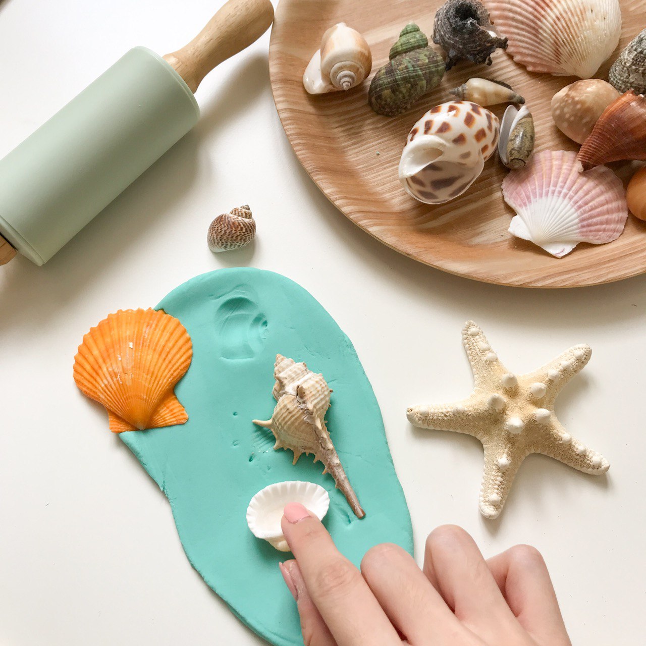 A Hand pressing a seashell into playdough to make an imprint. There are other assorted seashells in a brown plate and also a mint silicone roller on the side 