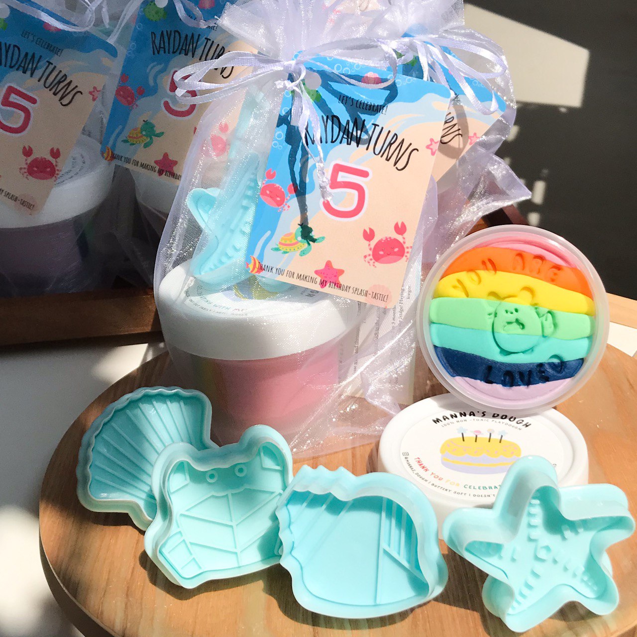 A party pack containing rainbow colored playdough, and various sea themed playdough cutters and neatly packaged in a organza bag with a ocean-themed gift tag design.