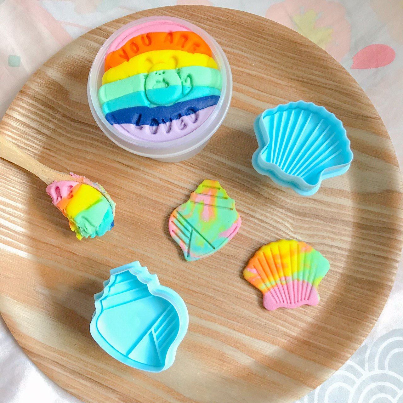 A rainbow tub of playdough along with two seashell playdough cutters and their rainbow playdough creations
